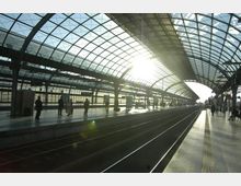 Überdachter Bahnhof mit moderner, gewölbter Glas- und Stahlkonstruktion. Personen stehen auf den Bahnsteigen, während Sonnenlicht durch das Dach einfällt und die Gleise erhellt.