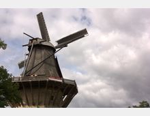 Windmühle mit hölzernem Aufbau und großen Flügeln auf einem steinernen Sockel, umgeben von Wolken am Himmel und einigen Bäumen im Vordergrund. Auf der Galerie der Mühle befinden sich einzelne Personen.
