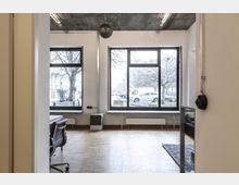 Bürobereich mit großen Fenstern, die Blick auf eine Straße mit Autos und kahlen Bäumen bieten. Innen sind Parkettboden, eine Bürostuhl-Ecke und moderne Lampen an der Betondecke sichtbar.