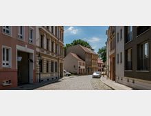 Kopfsteinpflasterstraße in einer Altstadt mit historischen Gebäuden, die teils mit dekorativen Fassaden und roten Ziegeldächern gestaltet sind. Im Hintergrund stehen Bäume, und parkende Autos sind am Straßenrand sichtbar.
