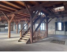 Offener Innenraum eines alten Backstein-Gebäudes mit Holzbalkendecke, Holzstützen und einer Holztreppe. Große Fenster mit Rundbögen lassen Tageslicht in den Raum, der eine Mischung aus rustikalem und industriellem Charme vermittelt.