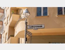 Straßenschild mit der Aufschrift „Schillerpromenade“ an einer historischen Straßenlaterne, im Hintergrund eine beigen Wohnhausfassade mit Balkonen und Fenstern.