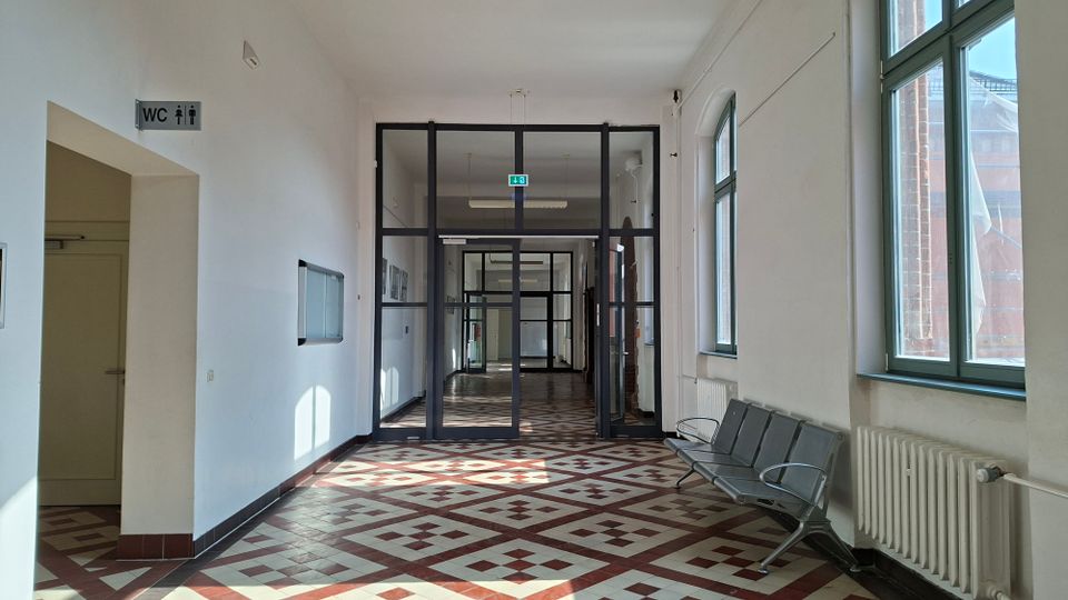Heller Innenflur in einem öffentlichen Gebäude mit weiß gestrichenen Wänden, großen Fenstern rechts und einer Sitzbank neben einem Heizkörper. Der Boden ist mit rot-weiß gemusterten Fliesen ausgelegt, und am Ende des Gangs führen dunkle Glasrahmentüren in weitere Korridore; links weist ein WC-Schild auf einen Nebenraum.