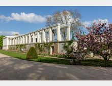 Eine historische Gartenanlage mit einer säulenartigen Pergola aus weißem Stein, umgeben von blühenden Magnolienbäumen und bunten Blumenbeeten. Im Hintergrund stehen große, kahle Bäume unter einem wolkigen blauen Himmel.
