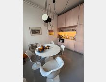 Modern open-plan kitchen and dining area with pale pink built-in cabinets and a lit backsplash along one wall. A round white dining table with white pedestal chairs sits under a large hanging pendant light, with framed artwork on the white walls and a gray floor.