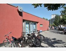 Flacher Gebäudeeingang mit einer rot gekachelten Fassade und einem stilisierten "U"-Symbol am oberen Rand, umgeben von Fahrradständern mit geparkten Fahrrädern; im Hintergrund sind weitere Gebäude sowie Bäume sichtbar.