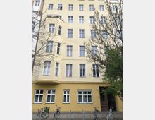 Fassade eines mehrstöckigen Wohnhauses mit gelber Putzfassade und weißen Fenstern. Im Erdgeschoss befinden sich ebenfalls Fenster sowie ein Hauseingang rechts, vor dem Gebäude stehen einige Fahrräder und Bäume mit kahlen Ästen.