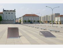 Weitflächiger, gepflasterter Platz mit Skateboarding-Rampen im Vordergrund, flankiert von historischen Gebäuden mit weißen Fassaden und roten Dächern. Im Hintergrund moderne Wohngebäude und vereinzelte Straßenlaternen.