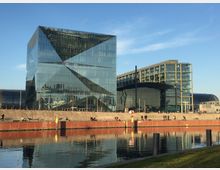 Moderne Glasgebäude am Wasser, bestehend aus einem geometrisch gestalteten, reflektierenden Würfel und einer angrenzenden rechteckigen Struktur mit großen Glasflächen. Im Vordergrund eine breite Promenade am Ufer und ein ruhiger Fluss, der die Gebäude spiegelt.