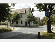 Eine Straßenszene mit einer Kreuzung, umgeben von Bäumen und einem zweistöckigen Gebäude mit Satteldach, das als Gasthaus gekennzeichnet ist. Im Vordergrund befinden sich Verkehrsampeln, Straßenschilder und ein Fußgängerweg.