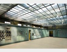 Raum mit industriellem Schaltanlagen-Design, gekennzeichnet durch eine Wand mit großen Kontrolltafeln, Schaltern und Anzeigen. Die Decke besteht aus einem Raster aus milchigen Glaspaneelen, und durch kleine Fenster in den Wänden fällt Tageslicht in den Raum.