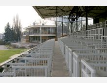 Ansicht einer überdachten Tribüne mit weißen Sitzgeländern und einem Blick auf ein modernes Gebäude mit Glasfassade im Hintergrund. Der Bereich ist von Grünflächen und Bäumen umgeben, mit einem gepflasterten Weg im Vordergrund.