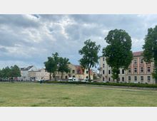 Eine städtische Szene mit historischen Gebäuden in Pastellfarben und roten Dächern, die eine Straßenfront bilden, flankiert von Bäumen im Vordergrund. Im Vordergrund befindet sich eine große Rasenfläche, während am Straßenrand Autos und ein Transporter parken.