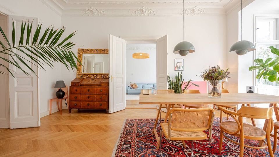 Helles Esszimmer in einer Altbauwohnung mit Stuckdecke, Fischgrätparkett und großem Holztisch auf gemustertem Teppich, umgeben von Stühlen und Hängelampen. Links steht eine antike Kommode mit Spiegel, rechts große Fenster und Zimmerpflanzen; durch offene Doppeltüren ist ein angrenzender Wohnraum sichtbar.
