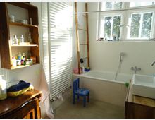 Helles Badezimmer mit weiß gefliester Badewanne, rechteckigem Waschbecken auf einem Holzunterschrank und einem großen Fenster mit Baumblick. Zur Ausstattung gehören ein weißer Heizkörper, ein Holzregal mit Pflegeprodukten und ein kleiner blauer Kinderstuhl.