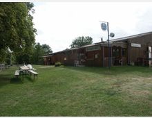 Holzgebäude mit der Beschriftung „Strandbad Imbiss” und „Biergarten” in ländlicher Umgebung, umgeben von Rasenflächen und Bäumen; davor stehen Bierbänke und Tische im Freien.