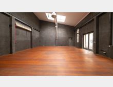 Industrie-innenraum mit hohen Decken, dunklen Wänden und einem großen Holzboden. Oberlichter, große Fenster und eine Glastür lassen Tageslicht einfallen, während die sichtbaren Rohre und Streben den industriellen Charakter unterstreichen.