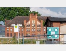 Historisches Backsteingebäude mit Giebelfront und zahlreichen Fenstern, im Vordergrund ein Bushaltestellenschild und ein grünes Notfallsammelplatz-Schild; im Hintergrund Bäume und bewölkter Himmel.
