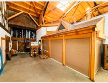 Innenansicht einer großen Lager- oder Werkhalle mit hoher Holzbinder-Dachkonstruktion und Oberlichtfenstern. In der Halle stehen eingebaute Holzboxen mit Rolltoren, daneben Betonboden sowie Regale und gelagerte Gegenstände entlang der Wände.