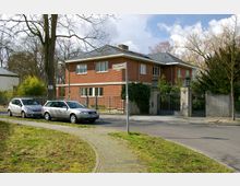 Zweistöckiges Wohnhaus aus roten Ziegelsteinen mit Flachdach, umgeben von einem Zaun und einer Einfahrt mit Tor. Vor dem Haus parken zwei silberfarbene Autos entlang einer Straße, die von Bäumen und Gehwegen gesäumt ist.