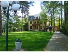 Historische Villa mit heller Fassade und Mansardendach, umgeben von einem großen Garten mit Rasen, Bäumen und gepflastertem Gehweg; im Vordergrund eine Laterne und ein Blumenkübel.