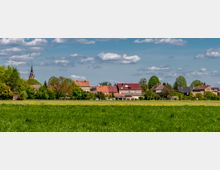 Eine ländliche Szene mit einer großen grünen Wiese im Vordergrund, dahinter eine Dorfansicht mit Häusern in unterschiedlichen Farben und Ziegeldächern. Links im Hintergrund ragt der Turm einer Kirche zwischen Bäumen hervor, auf den sich ein klarer blauer Himmel mit wenigen Wolken erstreckt.