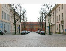 Kopfsteinpflasterstraße gesäumt von kahlen Bäumen und historischen Gebäuden in heller Putzfassade; am Ende der Straße steht ein großes rotes Gebäude mit weißen Fenstern und Satteldach. Geparkte Autos und einige Fußgänger sind sichtbar.