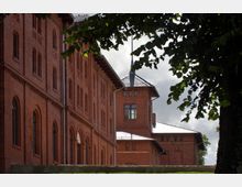 Backsteinbau mit mehreren Rundbogenfenstern und dekorativen Ziegelmustern, im Hintergrund ein Turm mit spitzem Dach. Der Bereich ist von Bäumen umrahmt und wird von einer grünen Rasenfläche begrenzt.