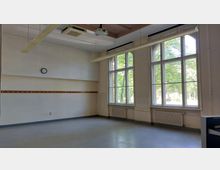 Klassenzimmer mit hohen Decken, einer Wand mit großen Fenstern, die den Blick auf Bäume und Tageslicht freigeben. Im Raum befinden sich eine Garderobenleiste, ein wandmontierter kleiner runder Wanduhr, eine Heizung sowie eine Projektionsvorrichtung an der Decke.