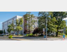 Wohnblock im urbanen Umfeld mit mehreren Etagen und gleichförmigen Fenstern, umgeben von Bäumen und einer grünen Fläche. Im Vordergrund sind eine Straßenkreuzung mit Verkehrsampeln und Schildern sichtbar, darunter ein Schild mit der Aufschrift "Rosa-Luxemburg-Straße".