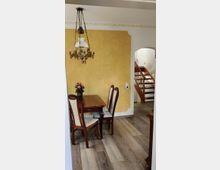 Esszimmer mit Holztisch und vier gepolsterten Stühlen, vor einer gelb gestrichenen Wand mit weißen Zierleisten. Eine dekorative Hängelampe hängt von der Decke, und im Hintergrund ist ein Treppenaufgang zu sehen.