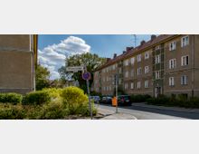 Wohnstraße mit mehrstöckigen Wohngebäuden aus hellem Putz und dunkelgrünen Details, daneben geparkte Autos. Ein Straßenschild mit der Aufschrift „Pfahlerstraße“ sowie Büsche und Bäume prägen den Vordergrund, bei leicht bewölktem Himmel.