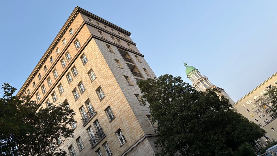 Schräg aufgenommene Ansicht eines hohen, rechteckigen Wohn- oder Bürogebäudes mit hellem Kachelmuster und vielen Fenstern, teils mit kleinen Balkonen. Im Vordergrund stehen große Bäume; rechts ragt hinter ihnen ein Turm mit grüner Kuppel vor blauem Himmel hervor.