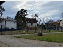 Wohngebiet mit Einfamilienhäusern, umgeben von Bäumen und Rasenflächen. Im Vordergrund ein eingezäuntes Grundstück, ein schmaler gepflasterter Gehweg und eine Rasenfläche mit vereinzelten Sitzbänken.