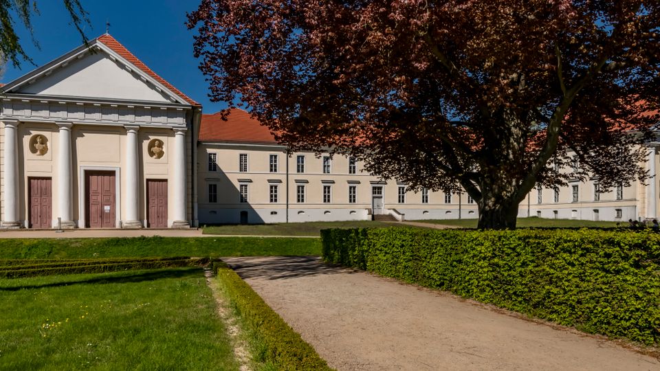 Wide view of a neoclassical white building with red-tiled roofs, featuring a columned entrance with three doors and a long rectangular wing with evenly spaced windows. A gravel path bordered by trimmed hedges and a large leafy tree leads through a grassy park setting in front of the building.