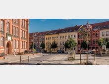 Historischer Stadtplatz mit gepflastertem Boden, flankiert von einem roten Backsteingebäude mit Rundbogenfenstern links und mehrfarbigen Gebäuden mit ornamentalen Fassaden im Hintergrund. In der Mitte stehen junge Bäume und ein Brunnen mit einer Statue.