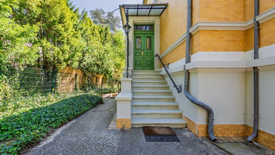 Seitlicher Eingangsbereich eines Backsteingebäudes mit einer Treppe zu einer grünen Doppeltür unter einem Glasvordach. Daneben verläuft ein gepflasterter Weg entlang eines Zauns und dichter Bepflanzung mit Bäumen und Bodendeckern.