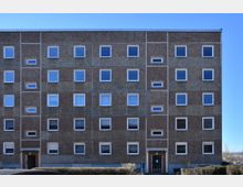 Ein viergeschossiges Plattenbau-Wohngebäude mit rechteckigen Fenstern in symmetrischer Anordnung auf der Fassade. Der Eingang ist im Erdgeschoss mittig platziert, umgeben von Hecken; der Himmel ist klar und blau.