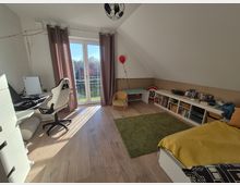 Ein modernes Kinderzimmer mit Dachschräge, großem Fenster und Terrassentür, die Tageslicht hereinlassen. Links steht ein Schreibtisch mit Computer, rechts ein Bett, Bücherregale und ein Tisch mit einem Stuhl; der Boden ist aus hellem Holz, teils mit grünem Teppich bedeckt.
