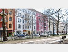 Straßenszene mit mehrstöckigen Altbauhäusern in verschiedenen Farben (Orange, Weiß, Rosa und Gelb), darunter befinden sich Geschäfte und Restaurants. Im Vordergrund befinden sich eine Allee mit kahlen Bäumen, ein Gehweg und eine Parkbank.