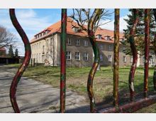 Ein historisches, mehrstöckiges Gebäude mit einem roten Ziegeldach und zahlreichen Fenstern, umgeben von einer teilweise begrünten Fläche, ist hinter einem bunt bemalten Metallzaun sichtbar. Im Vordergrund befinden sich kahle Baumäste, und im Hintergrund erstreckt sich ein klarer Himmel.