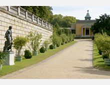 Ein Gartenweg, gesäumt von Pflanzkübeln mit gepflegten Sträuchern, führt zu einem gelben, klassizistischen Gebäude mit Säulenportikus und einer vergoldeten Figur auf dem Dach. Links verläuft eine hohe Steinmauer mit einer Bronzestatue davor.