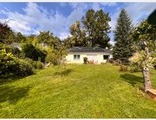 Großer Garten mit weitläufiger Rasenfläche vor einem eingeschossigen Bungalow mit hellem Anstrich und dunklem Dach mit Solarpaneelen. Dichte Hecken und hohe Bäume rahmen das Grundstück ein, daneben stehen Gartenmöbel und blühende Beete am Haus.