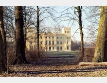 Helles, historisches Gebäude im klassizistischen Stil mit einem Turm, umgeben von kahlen Bäumen in einer parkähnlichen Landschaft. Der Vordergrund zeigt eine lichtdurchflutete Waldszene mit Laub am Boden.