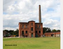 Backsteinindustriegebäude mit drei Etagen und hohem Schornstein, umgeben von einer großen Rasenfläche. Im Hintergrund sind weitere Gebäude und landwirtschaftliche Fahrzeuge zu sehen, bei bewölktem Himmel.