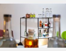 Ein Regal aus schwarzem Metall mit mehreren Ebenen, auf denen verschiedenfarbige Flaschen von Spirituosen und Getränken stehen, vor einer weißen Wand. Im Vordergrund sind unscharf weitere Flaschen sowie ein Teil eines grünen Sessels sichtbar.