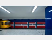U-Bahn-Station "Osloer Straße" in Berlin mit einer Wandgestaltung in den Farben der norwegischen Flagge (rot, blau und weiß). Links im Bild ist ein gelber U-Bahn-Zug in Bewegung zu sehen, während der Boden aus hellen Fliesen besteht und die Decke mit langen, leuchtenden Leuchtstoffröhren beleuchtet ist.