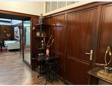 Innenraum mit dunkler Holzvertäfelung und Holzboden, daneben eine Glastrennwand und eine Tür. Im Vordergrund stehen Regal und Tische mit alten technischen Geräten und Messinstrumenten, im Hintergrund ist ein weiterer Raum mit Teppich und Möbeln zu sehen.