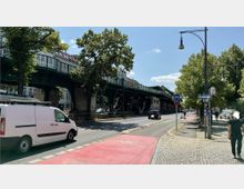 Straßenszene mit einer erhöhten, grünen Zugbrücke im Hintergrund, flankiert von Bäumen. Im Vordergrund sind eine Straße mit einem weißen Lieferwagen, ein Fahrradweg mit roter Markierung und ein gepflasterter Gehweg zu sehen, auf dem Passanten unterwegs sind.