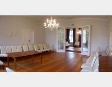 Ein heller, eleganter Raum mit Holzboden, weißen Wänden und cremefarben gepolsterten Stühlen, die entlang der Wände angeordnet sind. Im Zentrum steht ein Holztisch, und durch geöffnete Glastüren ist ein angrenzender Raum mit Vorhängen und weiteren Sitzgelegenheiten sichtbar.